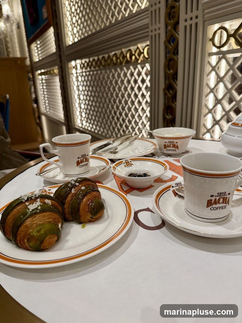 A table setting with two cups of coffee, two croissants, and a small bowl of jam. The cups and saucers are white with orange trim and feature the logo "1910 BACHA COFFEE". The croissants appear to be filled with a green cream and topped with nuts. The background shows decorative wall paneling with illuminated geometric patterns.