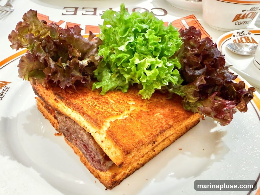 A close-up shot of a thick beef sandwich on toasted bread, topped with a generous portion of fresh green and red lettuce. The sandwich is served on a white plate with an orange rim, which also features embossed patterns. In the background, a white cup and saucer with the logo "1910 MARRAKECH BACHA COFFEE" are visible.