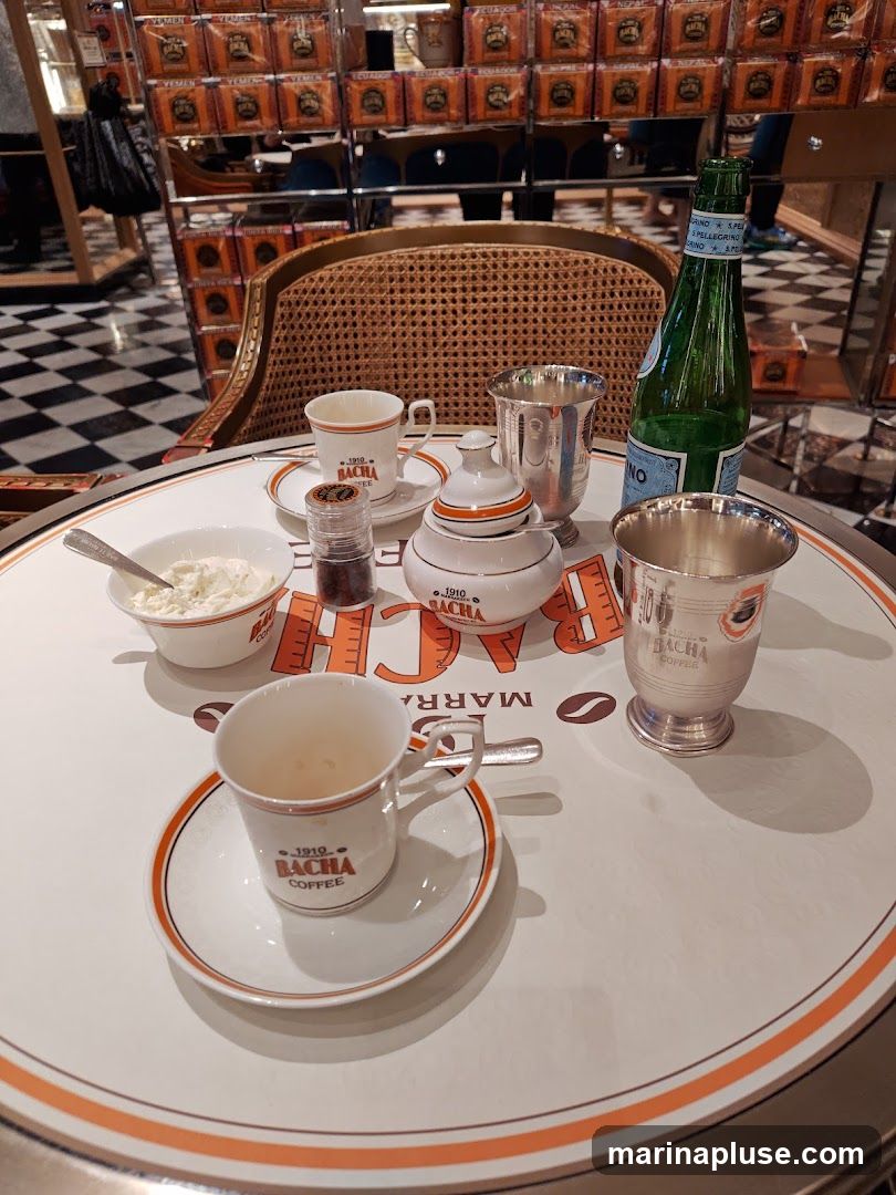 A close-up view of a round table set for coffee, featuring Bacha Coffee branding on cups, saucers, and the tabletop. The table is laid with two empty coffee cups and saucers, a sugar bowl, a small container of what appears to be sugar or spice, a bowl of cream, two silver cups, and a bottle of San Pellegrino water. In the background, shelves are stocked with numerous small boxes, likely containing coffee or tea, each labeled with country names such as 'Yemen' and 'Ecuador'. The flooring is a black and white checkered pattern.