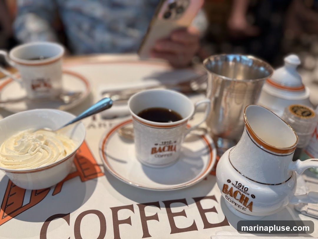A close-up shot of a table setting with coffee cups, a bowl of whipped cream, and a creamer. The cups and creamer are branded with "1910 MARRAKECH BACHA COFFEE". A person is visible in the background, holding a phone.