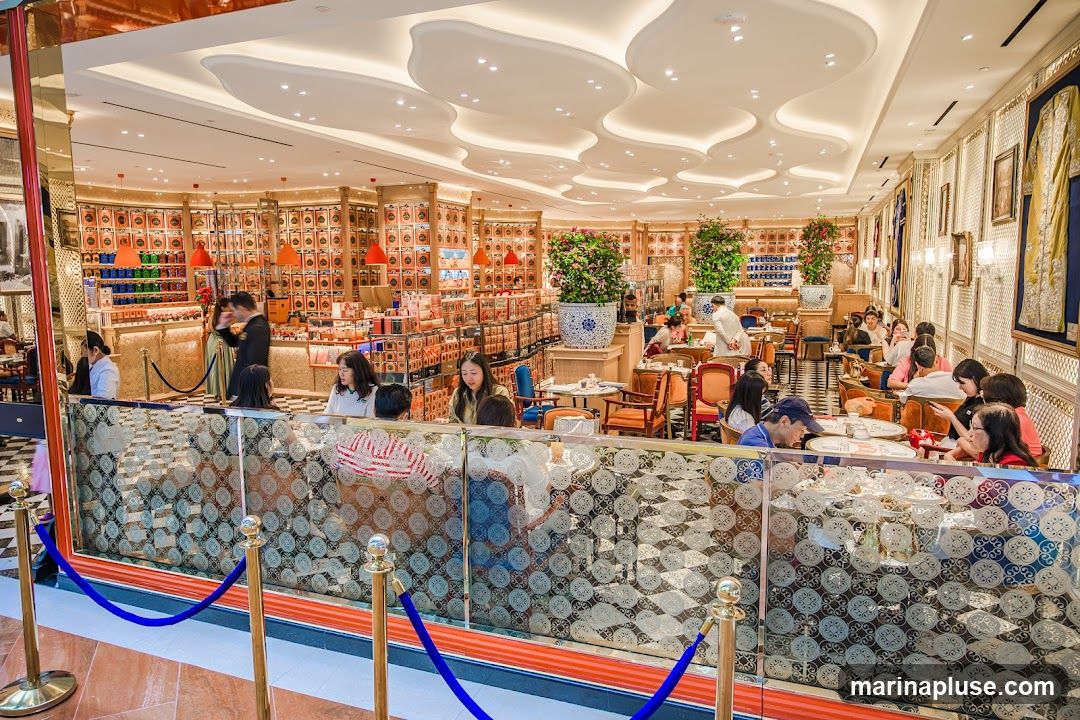 The image shows the interior of a cafe or restaurant with many patrons seated at tables. The walls are lined with shelves displaying numerous small boxes, possibly tea or other retail items. The decor is ornate, with decorative ceiling panels, patterned glass partitions, and elegant seating. There are also potted plants and framed artwork on display.