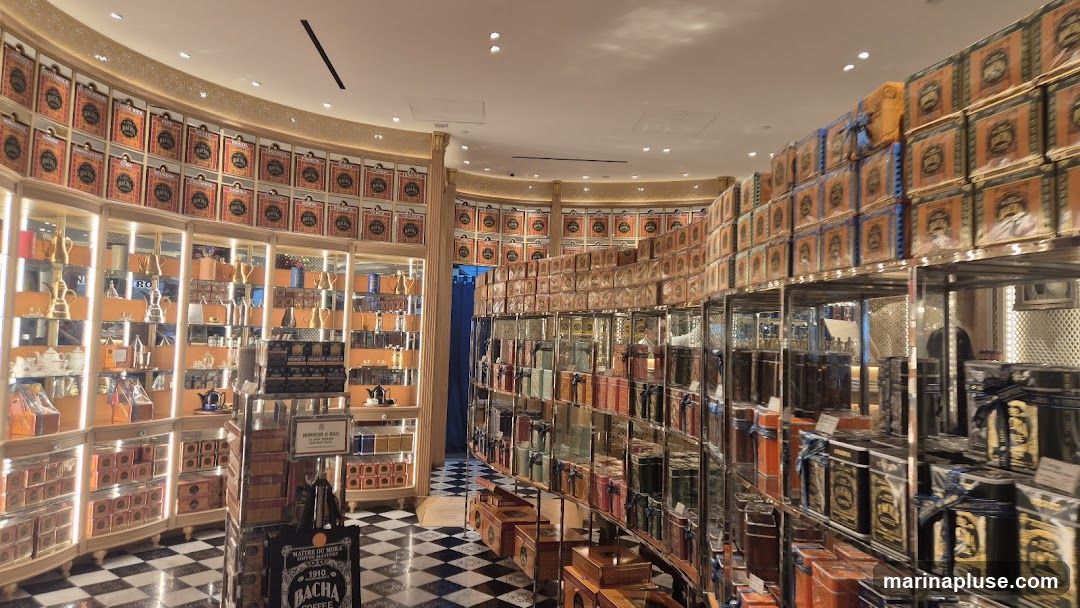The image shows the interior of a store, likely a tea or coffee shop, with numerous shelves filled with decorative tins. The tins are stacked high and arranged in curved rows, creating a visually striking display. Many of the tins feature ornate designs and appear to be branded. In the foreground, there are glass display cases showcasing various items, possibly teapots or other accessories. The floor has a distinctive black and white checkered pattern. The overall impression is one of abundance and a focus on high-quality, packaged goods.