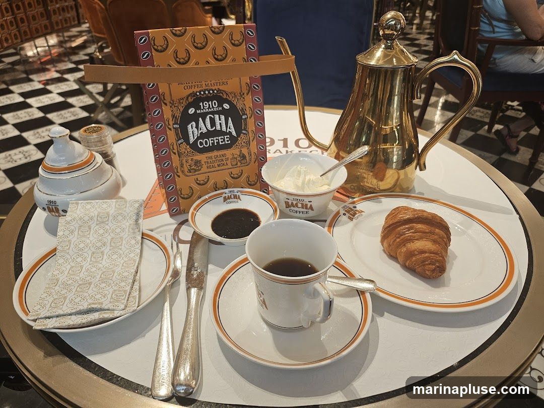 A table setting with a bag of Bacha Coffee, a golden coffee pot, a cup of coffee, a croissant on a plate, a small dish of dark liquid, a bowl of cream, a sugar dispenser, and a napkin. The Bacha Coffee bag is orange with a repeating pattern of crescent shapes and text. The coffee pot is ornate and golden. The cup and saucer are white with orange trim. The croissant is golden brown and flaky. The napkin is white with a repeating pattern.