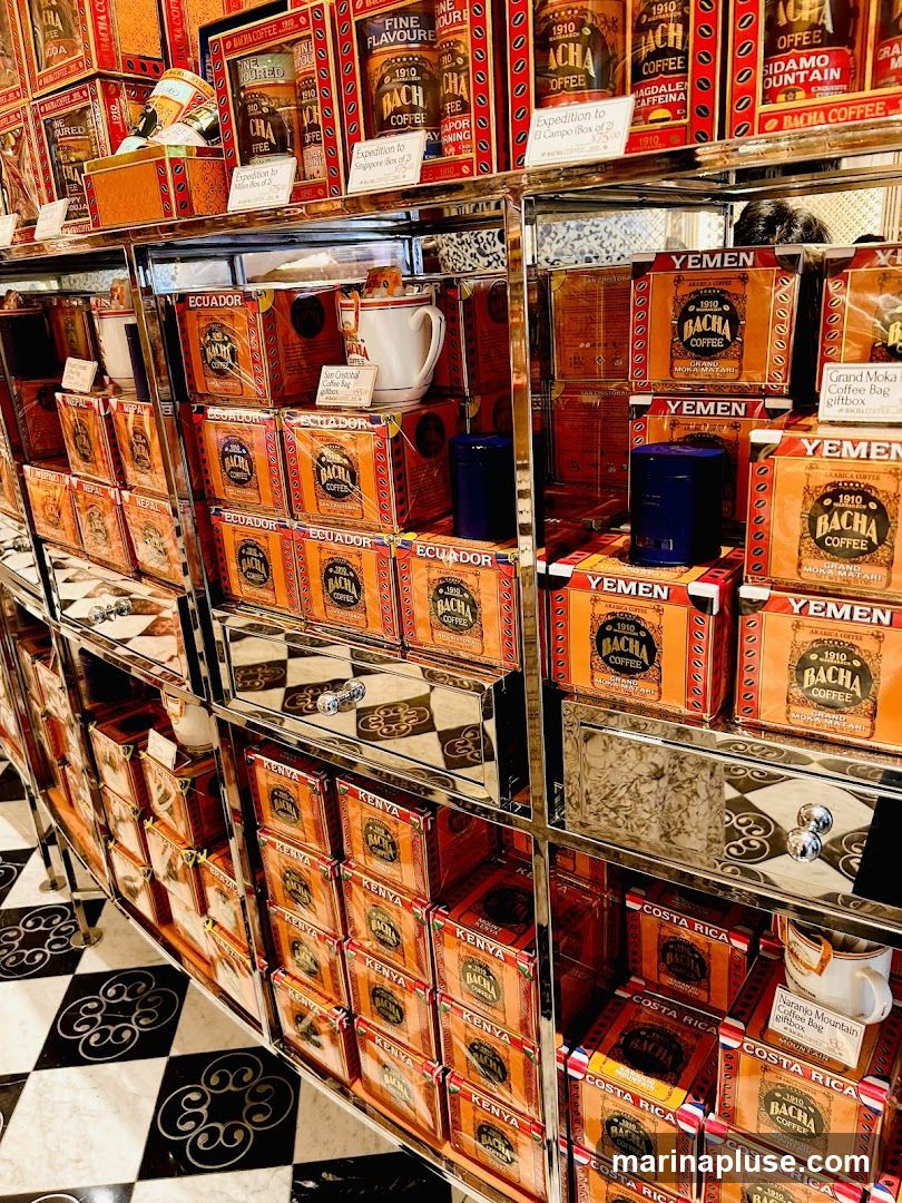 A retail display featuring numerous boxes of Bacha Coffee, organized by origin country such as Ecuador, Yemen, Kenya, and Costa Rica. The packaging is predominantly orange and gold with intricate designs. Some boxes are labeled as gift boxes or expeditions. A white mug with a tag is also visible. The shelves are made of chrome, and the floor has a black and white checkered pattern.