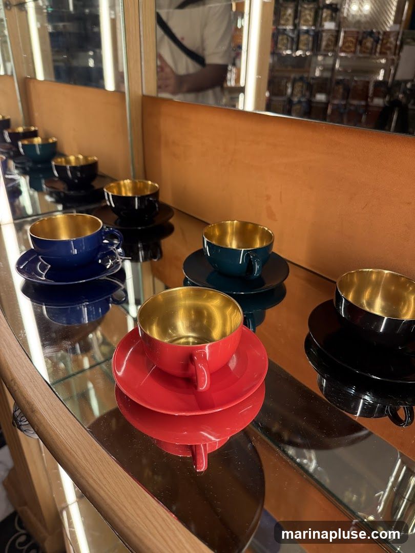 A display of colorful coffee cups and saucers with gold interiors is shown on a mirrored surface. The cups are arranged in rows, with colors including red, blue, black, and dark green. In the background, a person is visible in a mirror, and shelves stocked with items are also reflected.