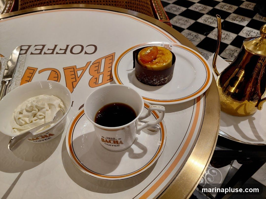 A table setting with a cup of black coffee, a small bowl of whipped cream, and a chocolate cake topped with caramelized fruit. A golden teapot is also visible. The table has a white surface with orange and gold stripes, and the brand name "BACHA COFFEE" is printed on the cup and saucer.