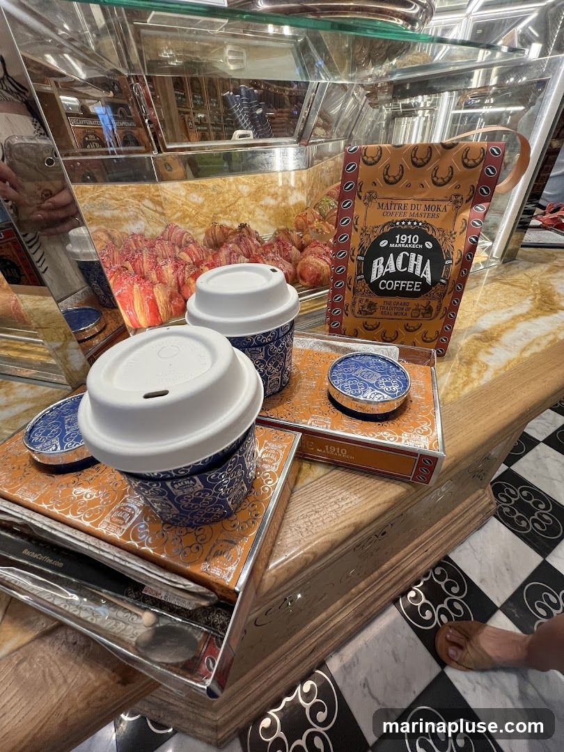 A display of coffee cups and pastries, with a bag of Bacha Coffee prominently featured. The coffee bag reads "Maitre du Moka Coffee Masters 1910 Marrakech Bacha Coffee The Grand Tradition of Real Moka Asia". Two coffee cups with white lids are on orange trays with blue and silver patterns. In the background, a glass display case shows rows of croissants and other pastries. The floor has a black and white checkered pattern.