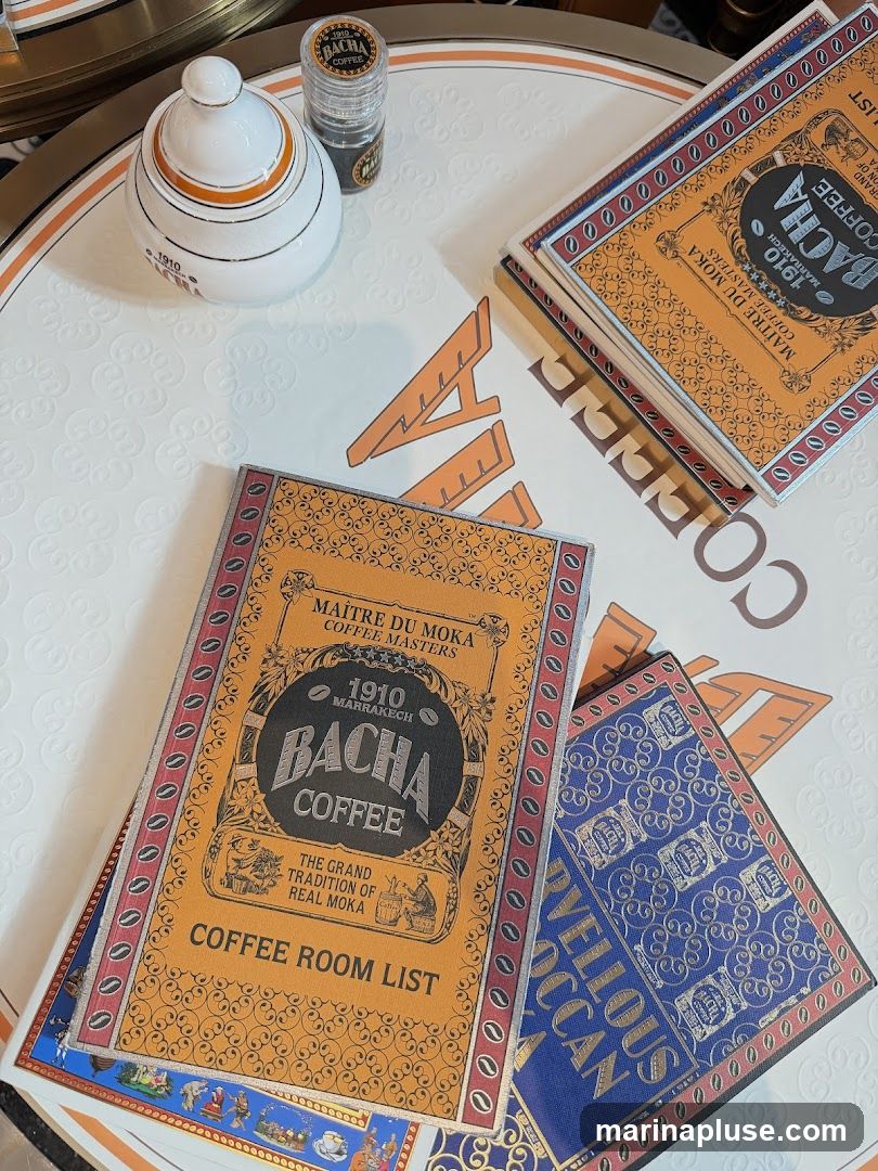A Bacha Coffee Room List menu is displayed on a table with other Bacha Coffee branded items, including a sugar bowl and a small jar. The menu is orange with ornate gold and red borders, featuring the Bacha Coffee logo and text.