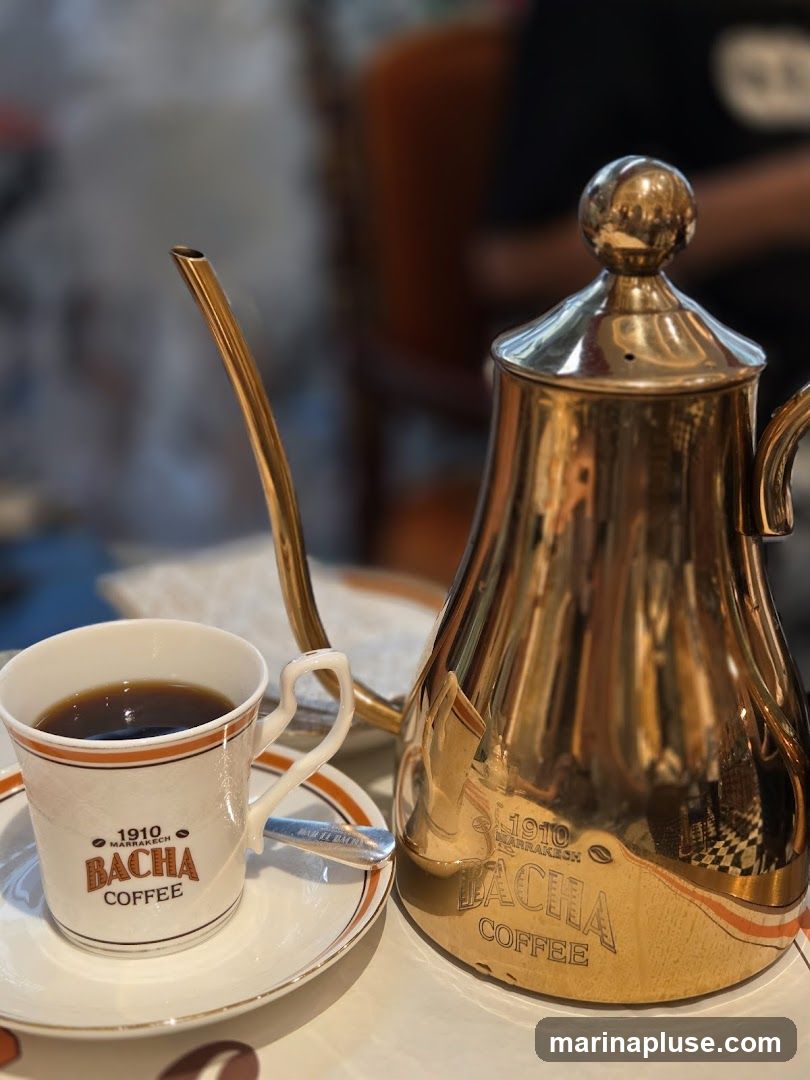 A close-up shot of a cup of coffee and a golden kettle, both branded with "1910 Marrakech Bacha Coffee". The coffee cup is white with orange trim and the Bacha Coffee logo. The kettle is ornate and reflective, also bearing the Bacha Coffee branding.