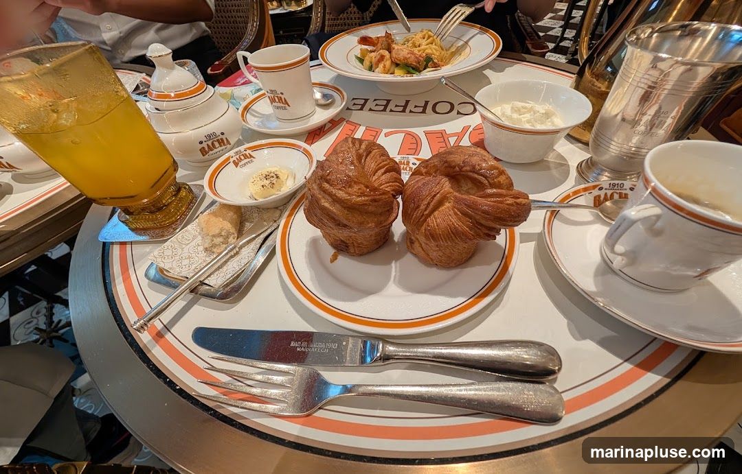 A table setting with two croissants, a bowl of butter, a bread roll, a glass of orange juice, a cup of coffee, and a plate of pasta with shrimp. The tableware and dishes are branded with "1910 Bacha Coffee".