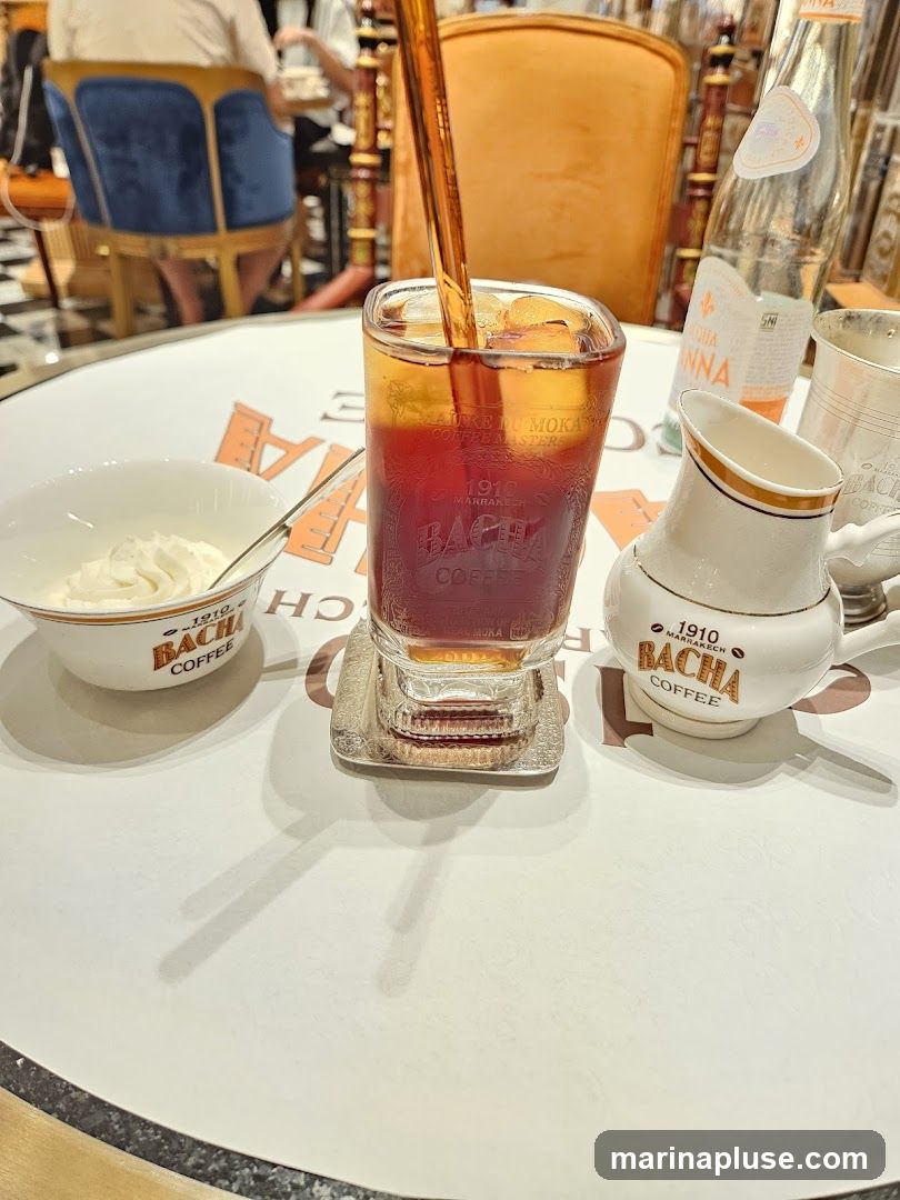 A close-up shot of an iced coffee drink being poured into a glass filled with ice. The glass has the branding "1910 MARRAKECH BACHA COFFEE" visible. To the left is a small bowl of whipped cream with the same branding. To the right is a small creamer pitcher, also branded "1910 MARRAKECH BACHA COFFEE". A bottle of San Pellegrino water is also on the table. In the background, blurred figures are seated at tables in what appears to be a cafe or restaurant.