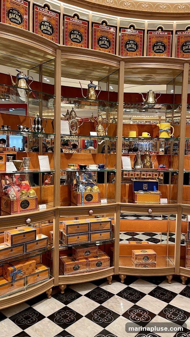 A display of coffee products and accessories in a shop. The shelves are filled with boxes of coffee, teapots, cups, and other decorative items. The walls are lined with framed labels for different coffee origins, such as Thailand, Hawaii, China, Zambia, and Vietnam. The floor has a black and white checkered pattern.