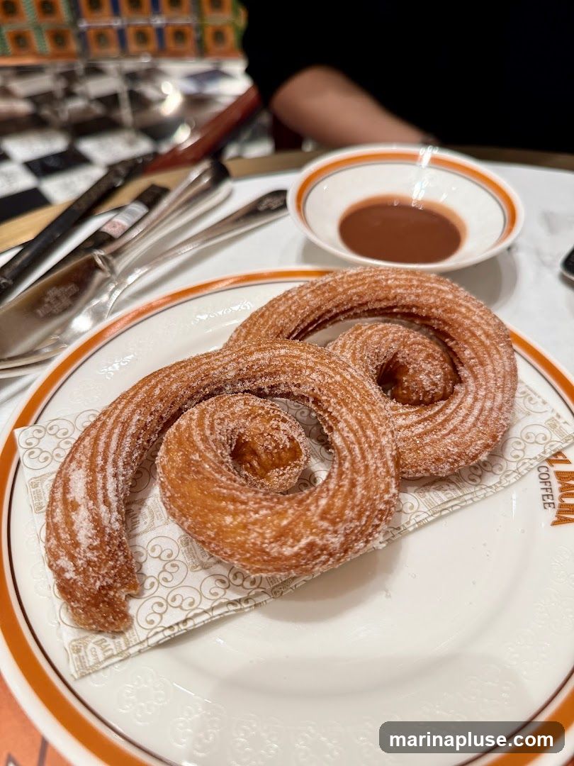 Two churros, coated in sugar, are served on a plate with a small bowl of chocolate dipping sauce. The plate and napkin have a logo that reads "MALIA COFFEE". The background shows a checkered pattern and cutlery.