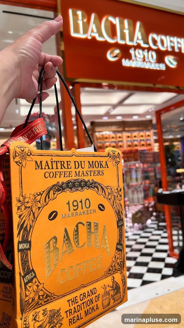 A hand holds an orange paper bag from Bacha Coffee, featuring ornate designs and text. In the background, the illuminated sign of a Bacha Coffee shop is visible, along with a checkered floor and shelves stocked with coffee products.