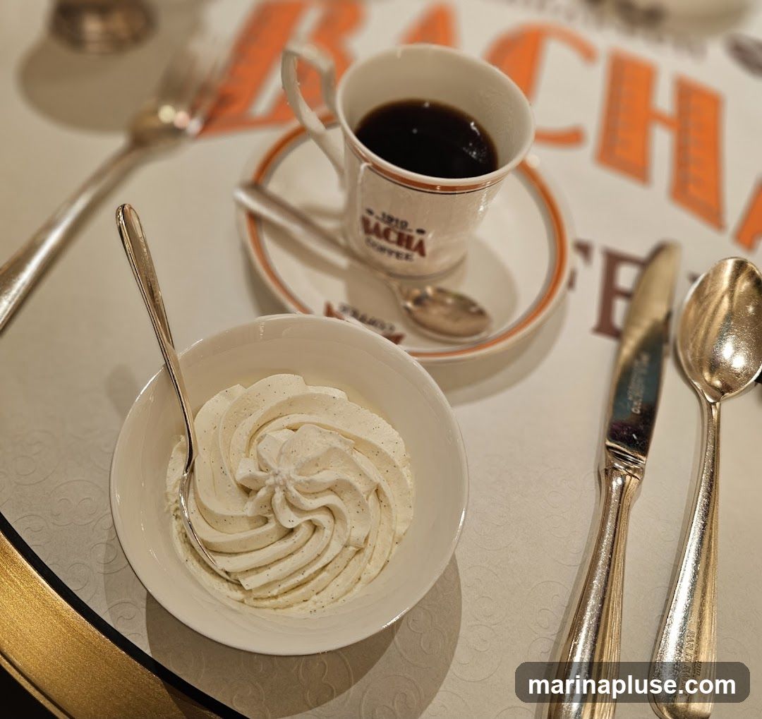 A close-up shot of a dessert and coffee set on a table. In the foreground, a white bowl is filled with whipped cream, swirled and garnished with vanilla specks. A spoon rests in the cream. Behind it, a white coffee cup with an orange rim sits on a saucer, filled with dark coffee. The cup has branding that reads "1910 SACHA COFFEE". A small spoon rests on the saucer. To the right of the coffee cup are a knife and spoon, both silver. The background shows a blurred orange and white pattern, possibly a tablecloth or placemat, with the word "SACHA" visible.