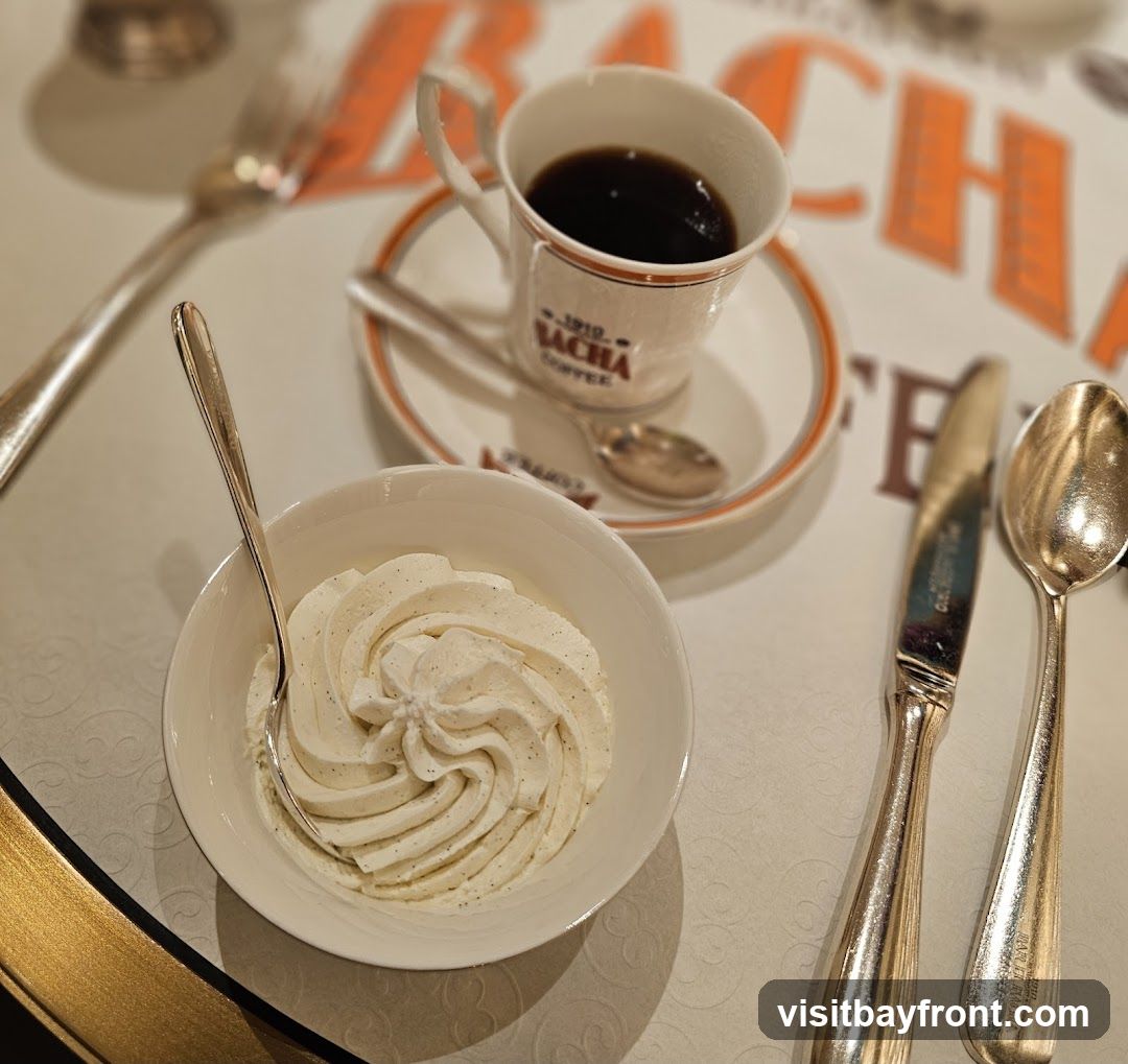 A close-up shot of a dessert and coffee set on a table. In the foreground, a white bowl is filled with whipped cream, swirled and garnished with vanilla specks. A spoon rests in the cream. Behind it, a white coffee cup with an orange rim sits on a saucer, filled with dark coffee. The cup has branding that reads "1910 SACHA COFFEE". A small spoon rests on the saucer. To the right of the coffee cup are a knife and spoon, both silver. The background shows a blurred orange and white pattern, possibly a tablecloth or placemat, with the word "SACHA" visible.