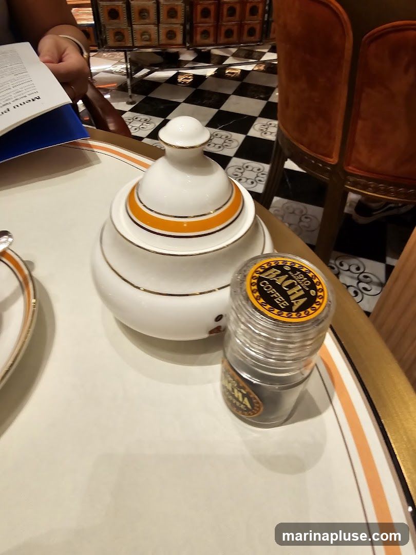 A close-up view of a table setting featuring a white sugar bowl with orange and gold trim, and a clear grinder containing dark coffee grounds. The grinder has a yellow label with black text that reads "BACHA COFFEE 1910". In the background, a checkered floor and an orange velvet chair are visible. A menu is partially visible on the left side of the image.