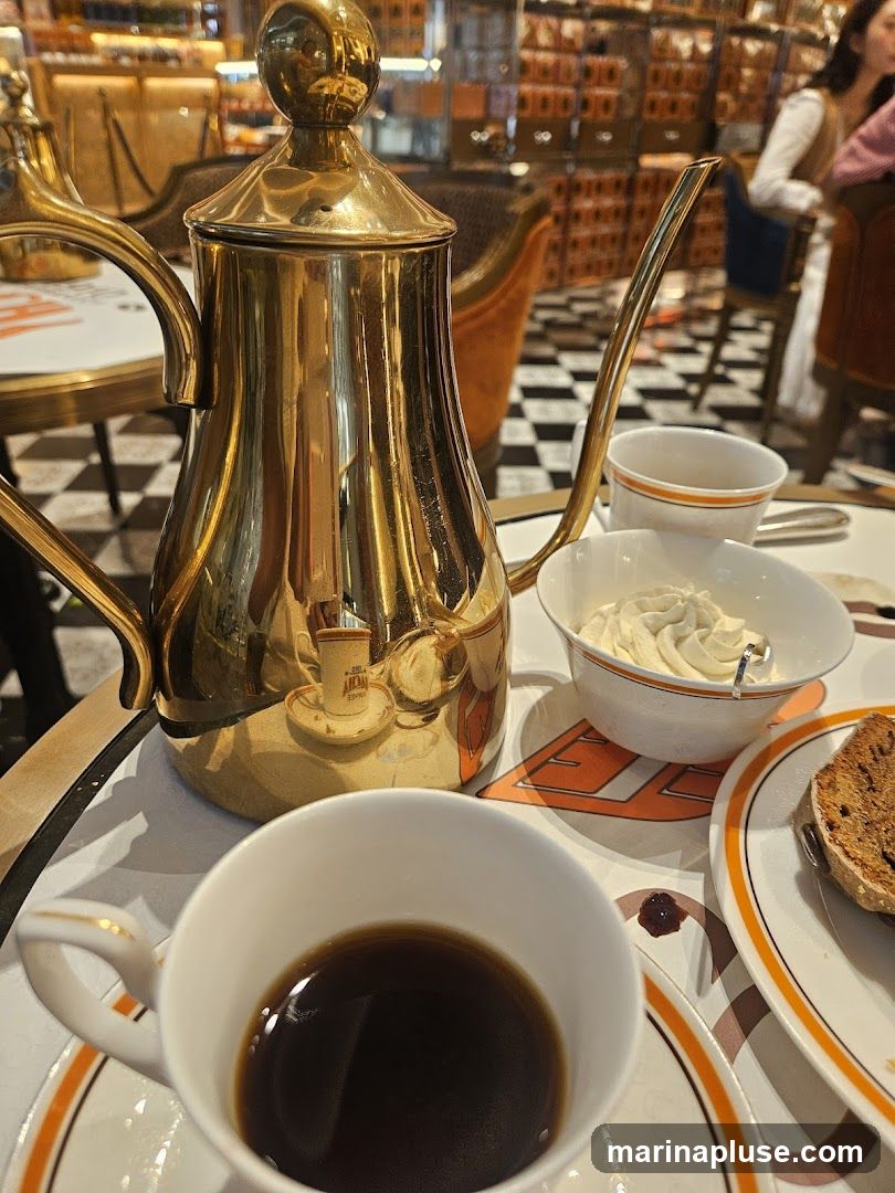 A close-up shot of a golden coffee pot, a cup of black coffee, a bowl of whipped cream, and a slice of pastry on a table. The background shows a blurred interior with checkered flooring and people dining.