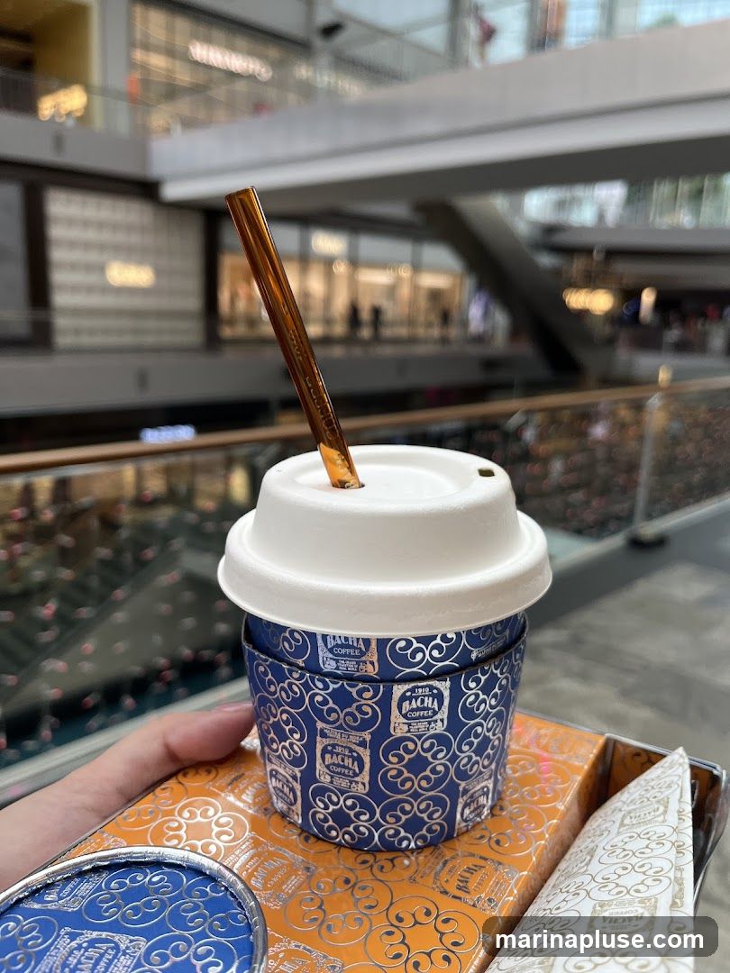A hand holds a coffee cup with a gold straw and a box, both decorated with a blue and silver pattern featuring the text "BACHA COFFEE". The background shows a blurred view of a shopping mall interior with shops on multiple levels.