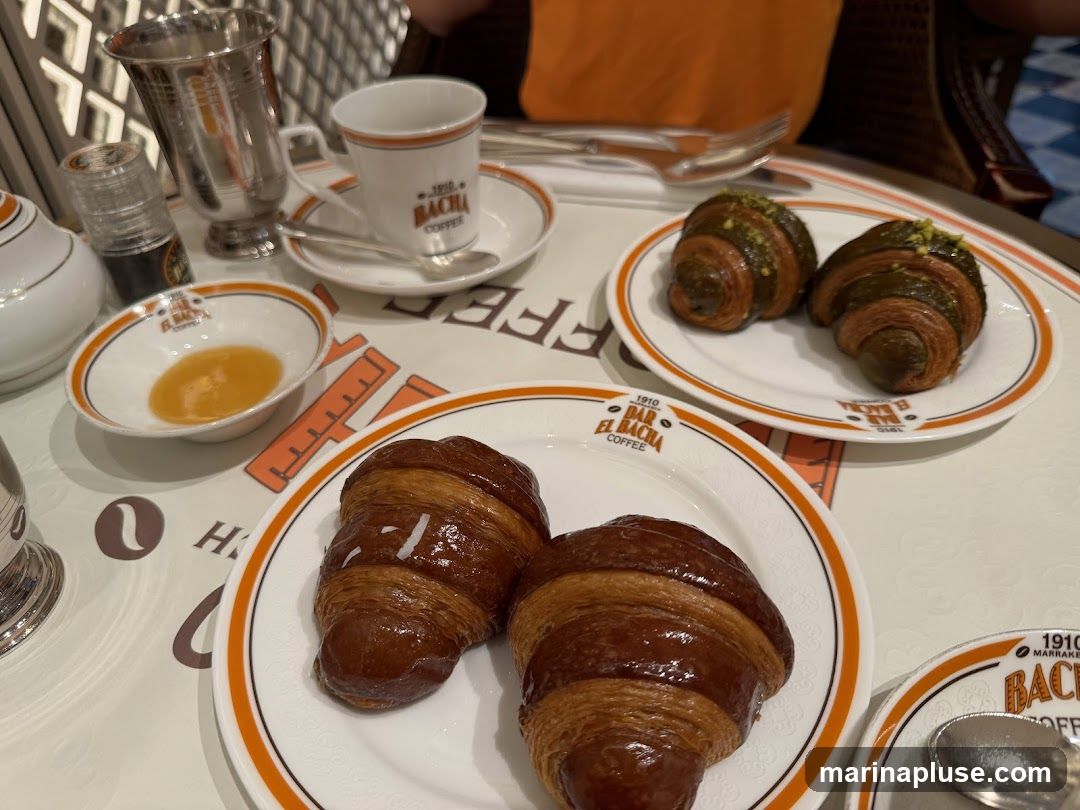 A close-up shot of a table setting with several croissants and a cup of coffee. Two plain croissants are on a white plate in the foreground. Behind them, on another plate, are two green-topped croissants. A small dish of honey and a coffee cup with a saucer are also visible. The branding "BAR EL BACHA COFFEE" is present on the plates and cup.