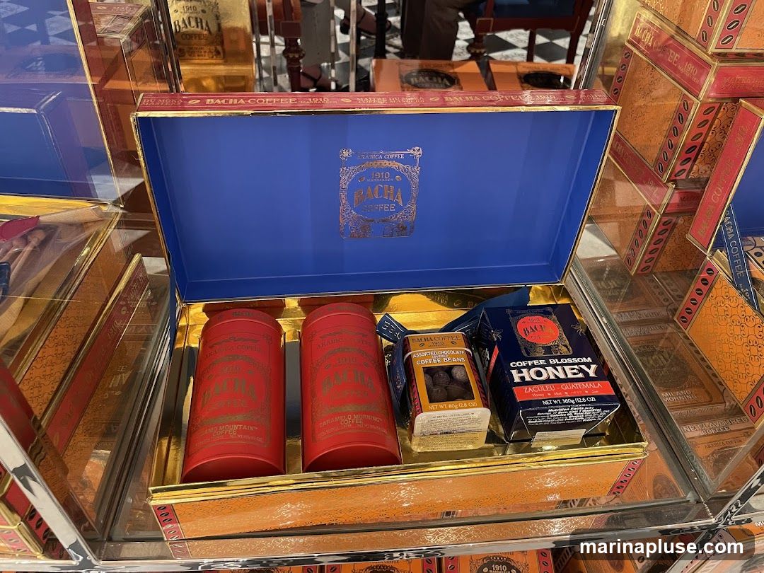 A Bacha Coffee gift box is open, revealing its contents. Inside are two red cylindrical containers of coffee, a small box of assorted chocolate coffee beans, and a box of coffee blossom honey from Guatemala. The box itself is gold with a blue interior, and the Bacha Coffee logo is visible on the lid. The gift box is surrounded by other Bacha Coffee products, suggesting a retail display.
