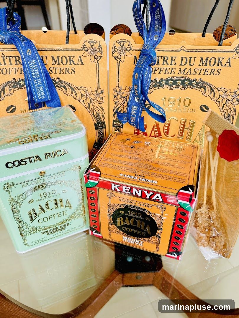 Three Bacha Coffee gift bags are visible, two with blue ribbons. In front of the bags are two tins of Bacha Coffee, one labeled "Costa Rica Volcan Azul" and the other "Kenya Mount Kenya". A bag of rock candy sticks is also present.