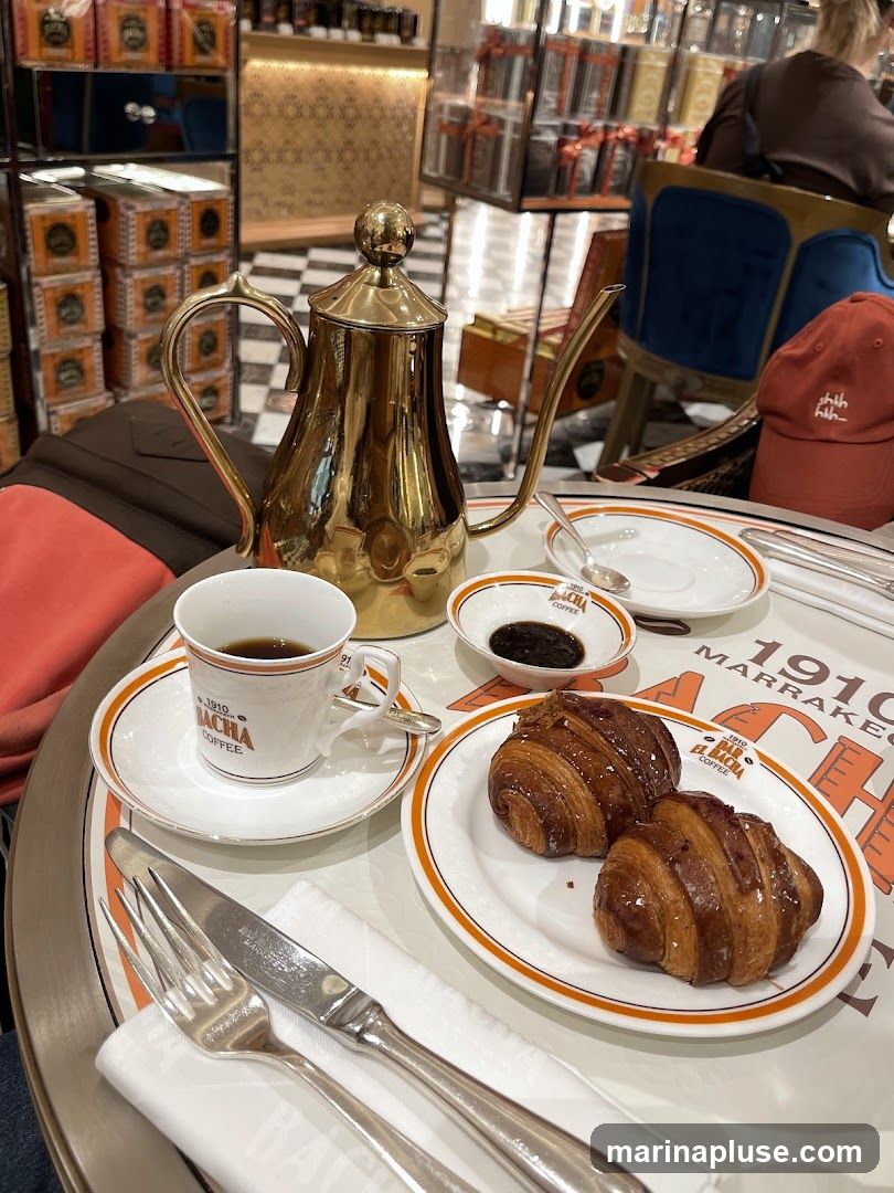 A close-up view of a coffee set with two croissants on a plate. The set includes a golden coffee pot, a cup and saucer with coffee, and a small dish with jam. The cup and saucer have the branding "1910 EL BACHA COFFEE". In the background, shelves are stocked with boxes, and a person is seated at a table.