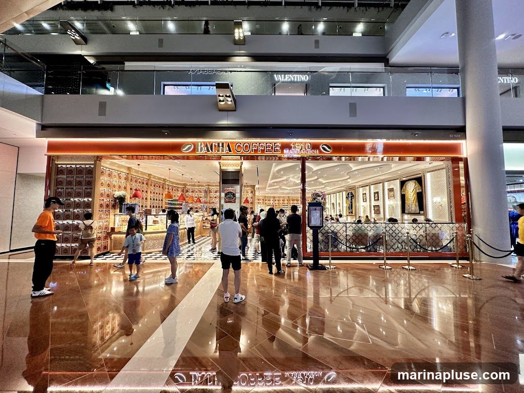 The exterior of a Bacha Coffee shop with its distinctive orange and gold branding. The shop has large glass windows and doors, allowing a view of the interior seating area and displays. The shop's name and logo are prominently displayed above the entrance. People are seen entering and exiting the shop, and some are standing outside. The floor in front of the shop is highly reflective, showing the shop's facade and the surrounding mall environment.