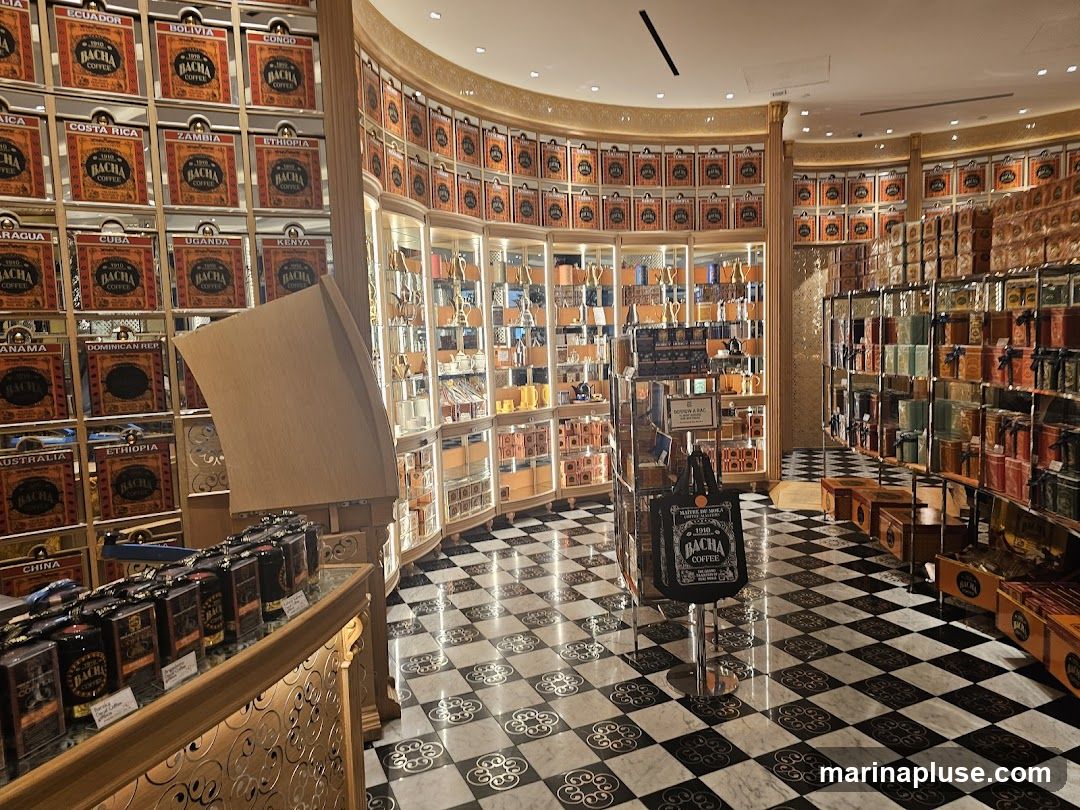 The interior of a coffee shop with walls lined with shelves displaying numerous coffee tins and boxes. The tins are labeled with country names and the brand "BACHA COFFEE". In the center, there is a display stand with a tote bag and some smaller boxes. The floor has a black and white checkered pattern with decorative elements. Glass display cases showcase more products and coffee-related items.