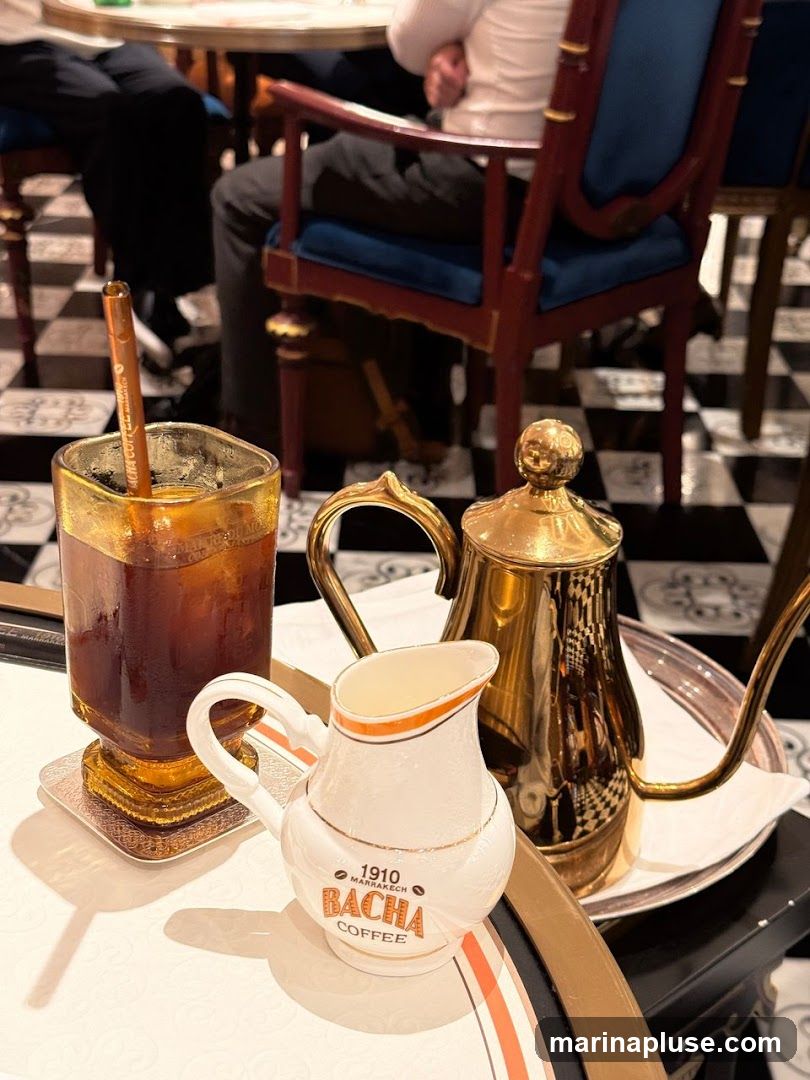 A close-up of a coffee service on a tray, featuring a glass of iced coffee, a small creamer pitcher with the Bacha Coffee logo, and a golden teapot. The setting appears to be a cafe or restaurant with checkered flooring and ornate chairs.