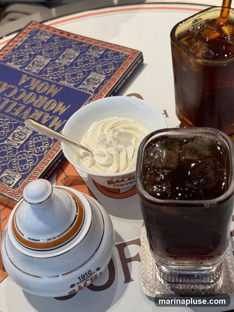 A close-up shot of a table setting featuring two glasses of iced coffee, a bowl of whipped cream, a sugar bowl, and a decorative book. The glasses and sugar bowl have branding that appears to be from a coffee establishment. The book is titled "MARVELLOUS MOKA MOROCCO".