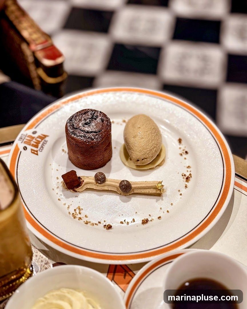 A close-up shot of a dessert on a white plate with an orange rim. The dessert consists of a chocolate roulade, a scoop of ice cream, and a decorative element with chocolate spheres and gold leaf. The plate has a logo that reads "BAR EL BACIA COFFEE 1915". In the background, a black and white checkered floor and a chair with red and gold accents are visible. A cup of coffee and a bowl of cream are also partially visible in the foreground.