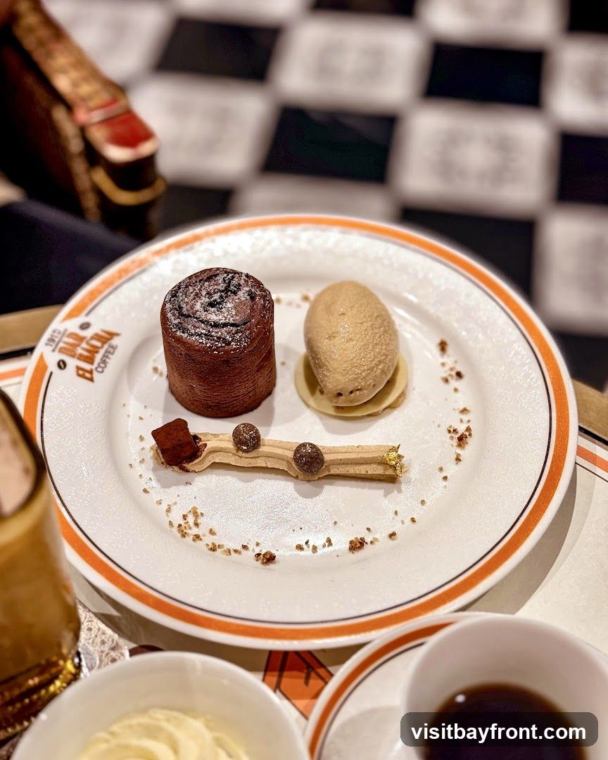 A close-up shot of a dessert on a white plate with an orange rim. The dessert consists of a chocolate roulade, a scoop of ice cream, and a decorative element with chocolate spheres and gold leaf. The plate has a logo that reads "BAR EL BACIA COFFEE 1915". In the background, a black and white checkered floor and a chair with red and gold accents are visible. A cup of coffee and a bowl of cream are also partially visible in the foreground.