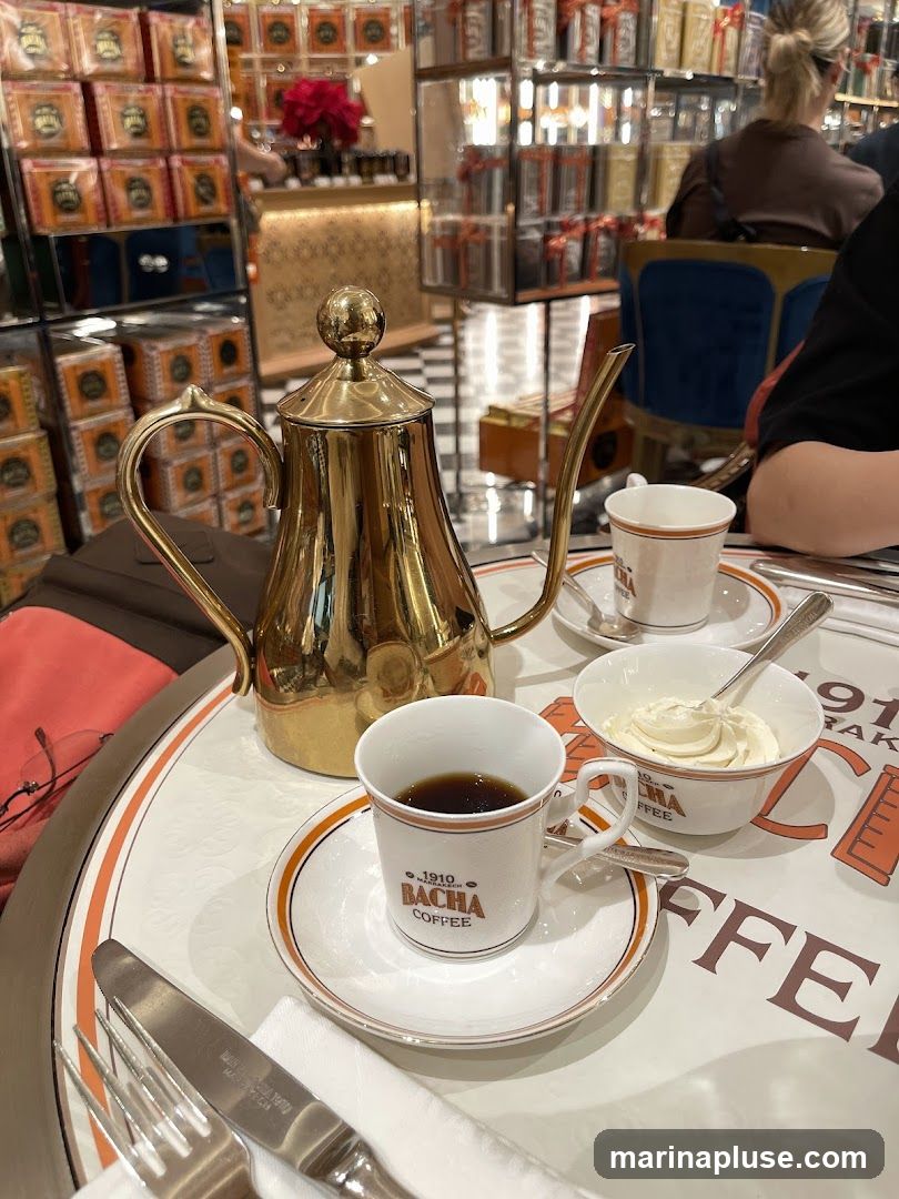 A golden teapot sits on a table with two cups of coffee, a bowl of cream, and silverware. The cups and bowl are branded with "BACHA COFFEE". In the background, shelves are filled with boxes of coffee and other products.