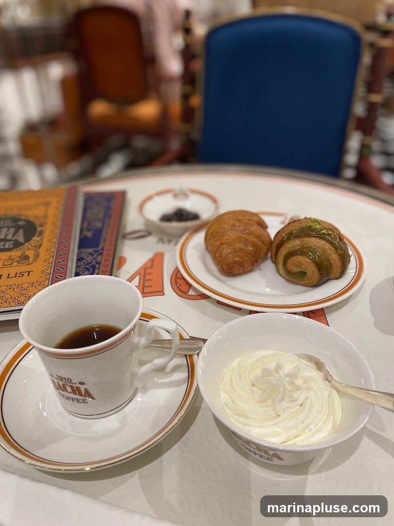 A close-up view of a coffee cup, saucer, and a bowl of whipped cream on a table. Two croissants are on a plate in the background. A book or menu is also visible on the left. The items appear to be from a coffee shop.