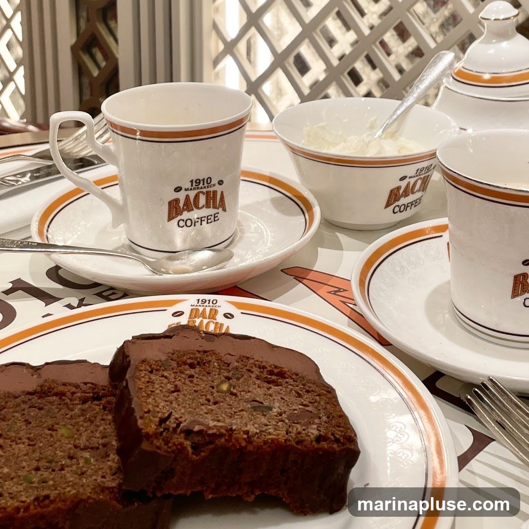 A close-up shot of two slices of chocolate cake with nuts and frosting, served on a white plate with orange trim. Two coffee cups and saucers, also with orange trim and branded with