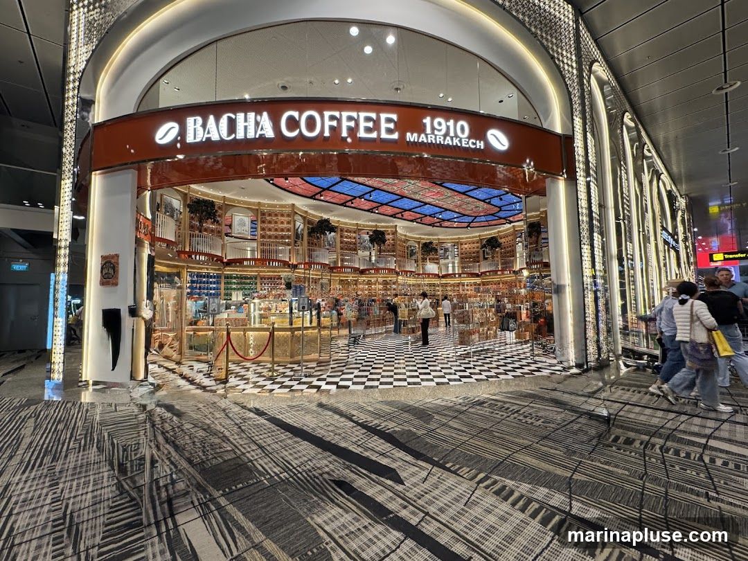 The exterior of a Bacha Coffee shop with its prominent signage. The shop features a distinctive checkered floor and displays various coffee-related products and merchandise. The interior is visible through the open entrance, showcasing a luxurious and ornate design with shelves stocked with goods.