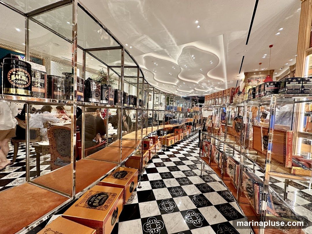 The image shows the interior of a store with a checkered black and white floor. Tall shelves line the walls, filled with various products, including large tins labeled "BACHA COFFEE" and smaller boxes labeled "HONEY". Some shelves also display what appear to be books or decorative boxes. In the background, people are seated at tables, suggesting a cafe or seating area within the store. The ceiling has an abstract, wavy design with recessed lighting. The overall aesthetic is luxurious and well-organized.