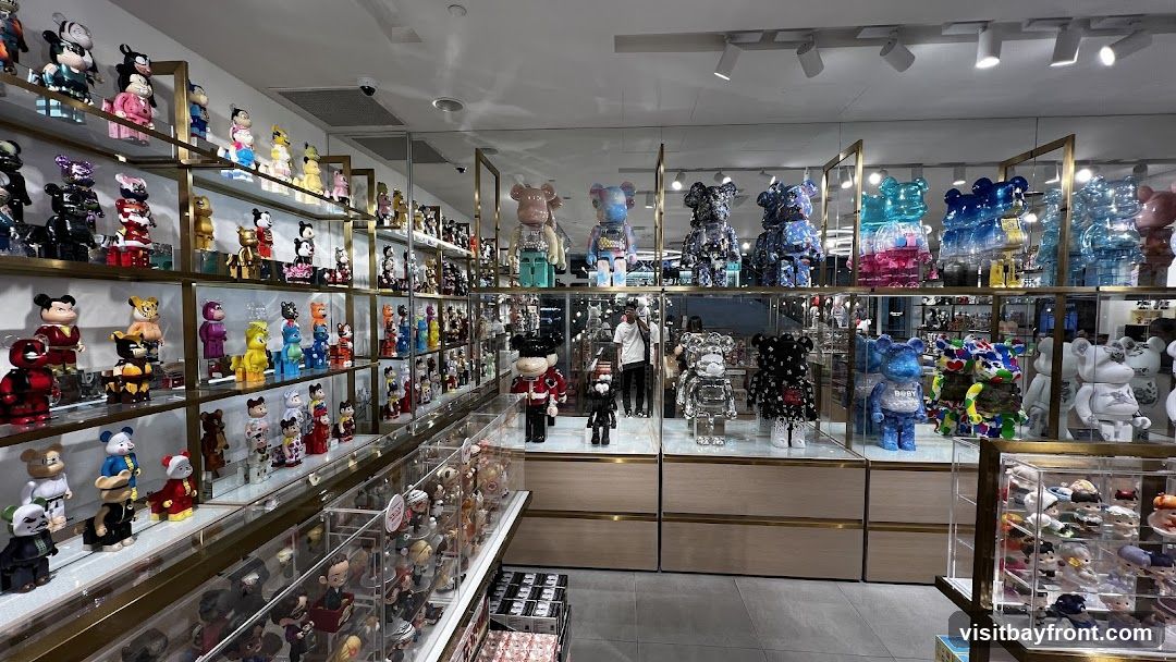 The interior of a store filled with display shelves showcasing a large collection of collectible toys, primarily Bearbrick figures in various sizes and designs. The toys are arranged on multiple levels of glass shelves, some enclosed in display cases. In the background, a person is visible standing near a display of larger figures. The store appears well-lit with overhead lighting.