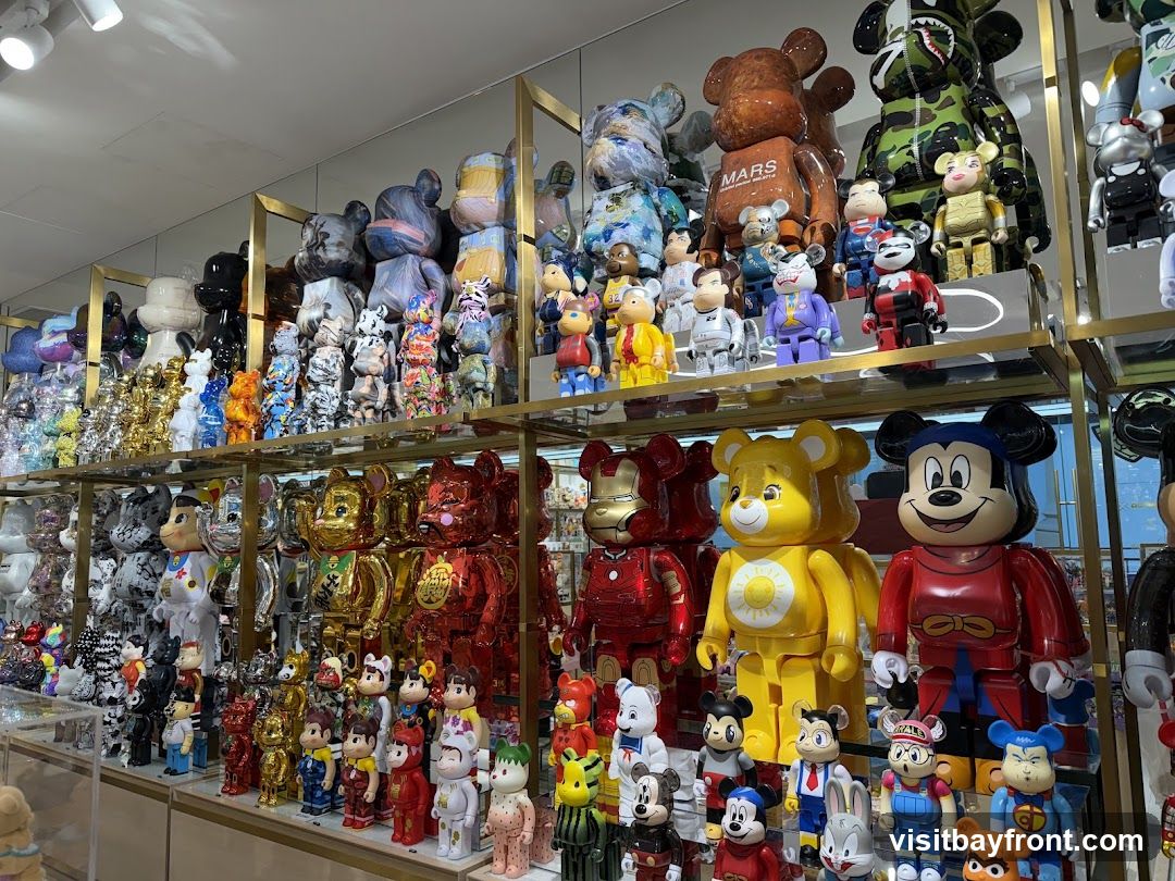 A retail display featuring a wide variety of collectible figures, primarily Bearbrick-style toys, arranged on multiple shelves. The figures come in various sizes, colors, and designs, including characters like Mickey Mouse, Iron Man, and Care Bears, as well as abstract patterns and camouflage prints. The shelves are illuminated, highlighting the glossy finishes of many of the figures.