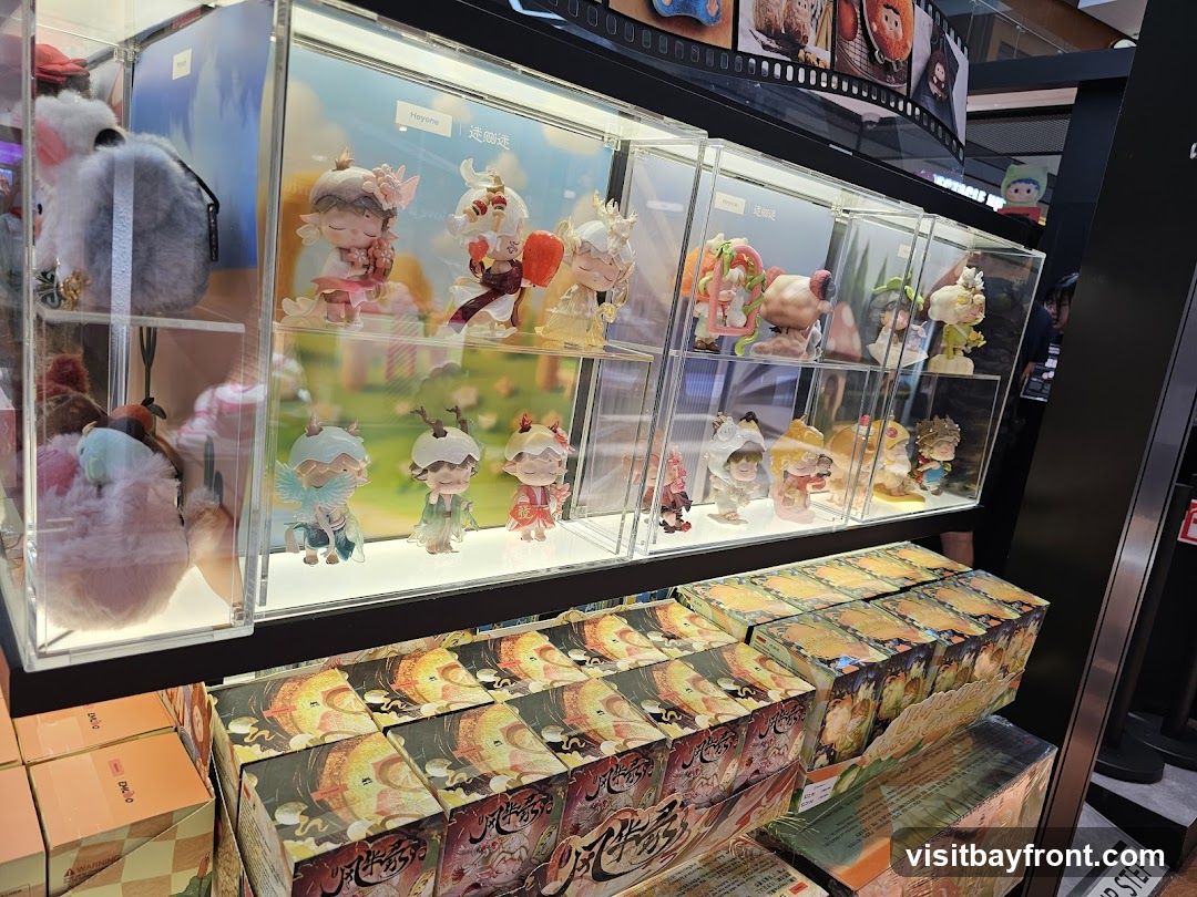 A retail display featuring several glass cases filled with collectible figurines. The figurines are varied in design, with some appearing to be part of a series. To the left, plush toys and bags are also displayed. Below the display cases, stacks of boxed merchandise are arranged, featuring intricate artwork. The overall impression is of a store selling character-based merchandise.