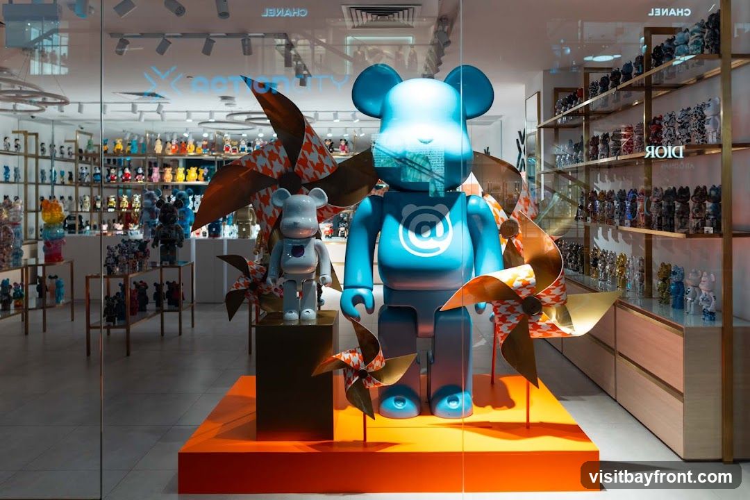 A store window display features two large blue bear-shaped figures, one significantly larger than the other, both adorned with an '@' symbol. The figures are surrounded by decorative pinwheels with an orange and white houndstooth pattern. Behind the display, shelves are filled with numerous smaller collectible bear figures in various colors and designs. The store interior is visible, showing more shelves and displays of merchandise. The reflection on the glass shows the word 'CHANEL' and 'DIOR' on shelves in the background.