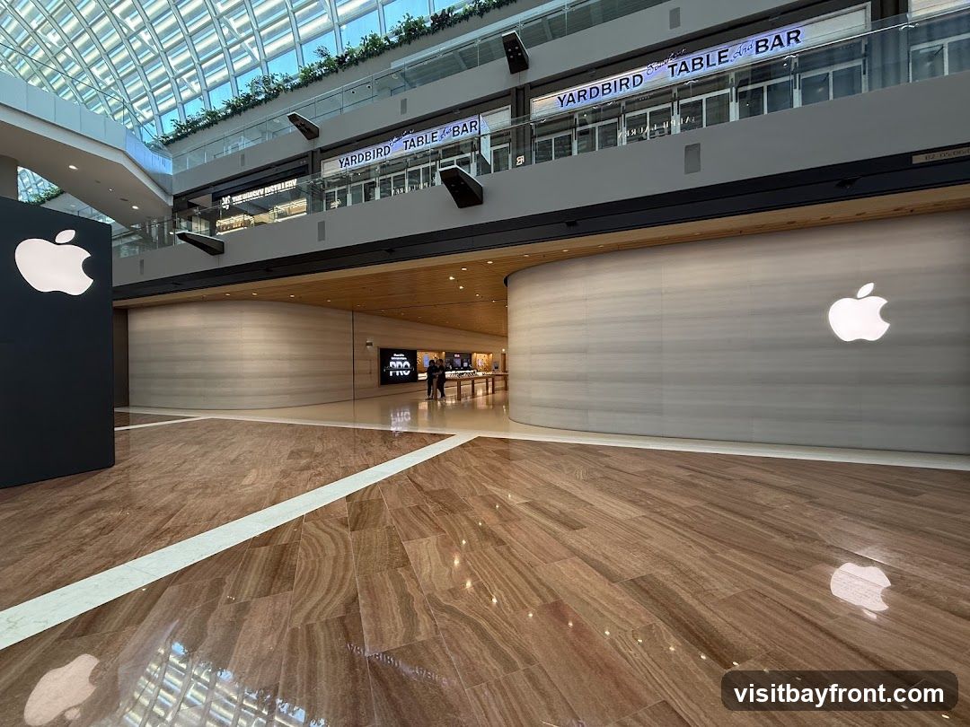 A wide shot of a modern shopping mall interior. In the foreground, a polished wooden floor reflects the overhead lighting and the Apple logo. To the left, a large black display features a prominent white Apple logo. In the background, the curved facade of an Apple Store is visible, with its iconic logo illuminated on the wall. Above the Apple Store, on an upper level, signage for "YARDBIRD TABLE And BAR" is displayed. The overall scene conveys a sense of contemporary retail space.
