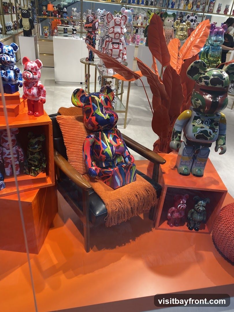A vibrant display featuring numerous collectible figurines, primarily of the Bearbrick design, in various colors and patterns. In the foreground, a large, multi-colored Bearbrick sits on a chair with an orange throw blanket. To its right, a Boba Fett-themed Bearbrick stands on an orange platform. Smaller figurines are visible on shelves and in cubbies throughout the display. The background shows a store interior with more shelves stocked with similar collectible items.