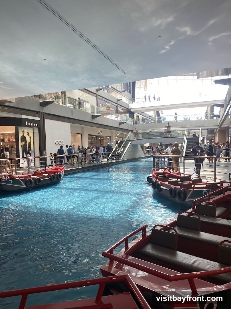 An indoor shopping mall features a canal with gondolas, surrounded by shops on multiple levels. The water is a vibrant blue, reflecting the light from the ceiling. People are seen walking along the balconies and stairs, and some are boarding the gondolas. Shops like Pedro and COS are visible.