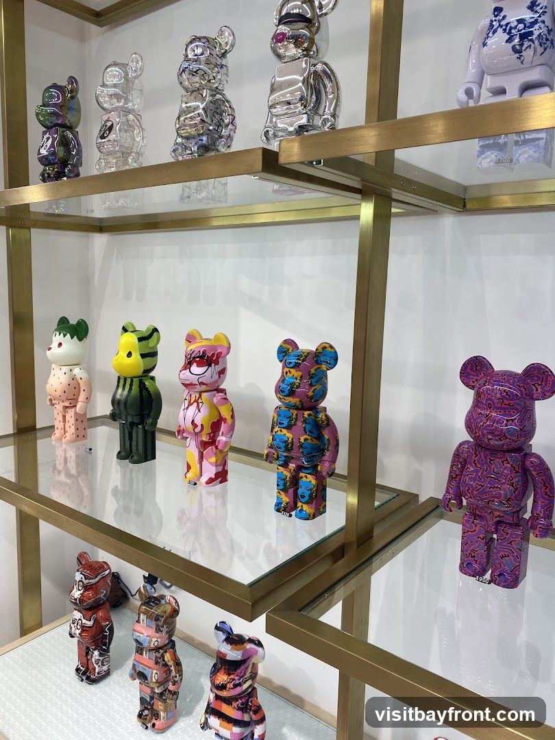 A retail display featuring multiple designer toys, primarily in the shape of stylized bears, arranged on glass shelves with gold-colored metal supports. The toys exhibit a variety of colors, patterns, and finishes, including metallic, iridescent, and graphic prints. Some toys have visible price tags.