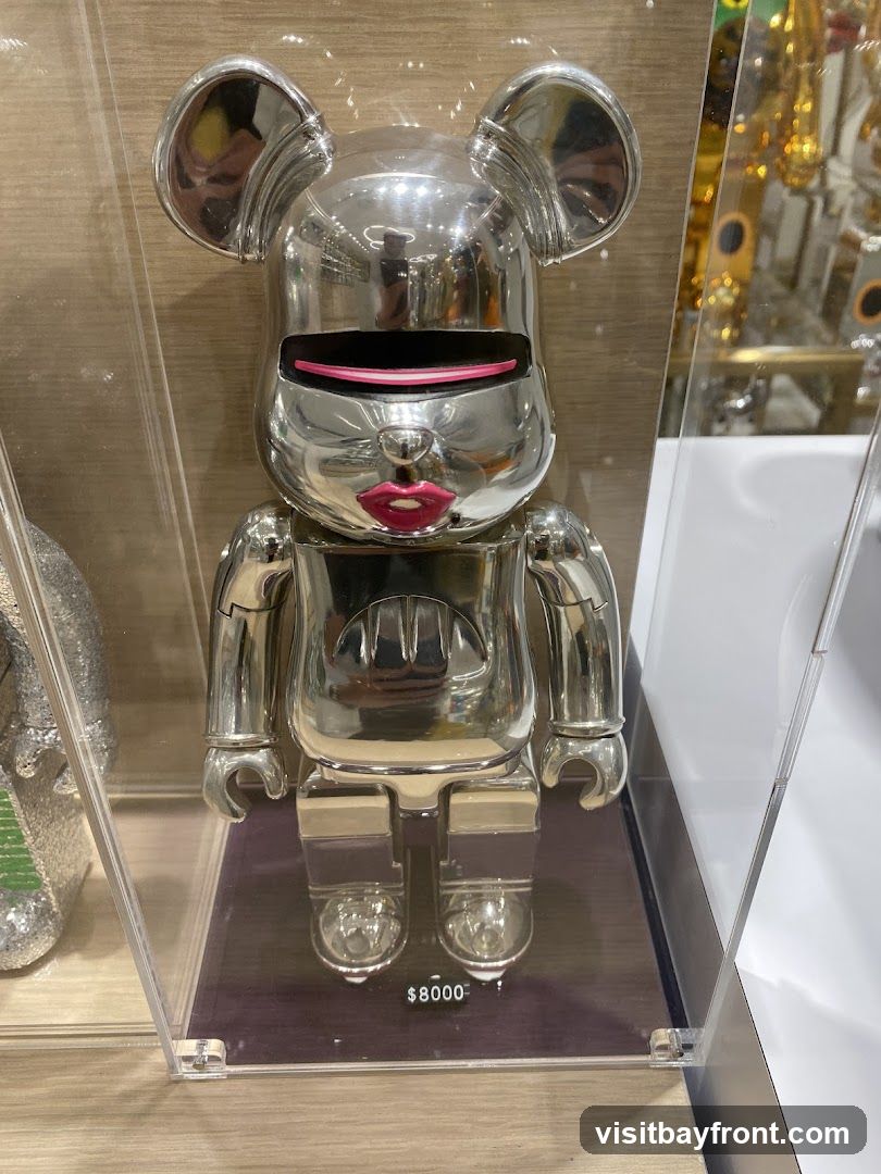 A shiny silver Bearbrick figure with pink lips and a black visor is displayed in a clear acrylic case. The figure has a stylized bear-like head with round ears and a robotic body. A price tag of $8000 is visible below the figure. The background appears to be a store interior with shelves and other items, some of which are also metallic and reflective.