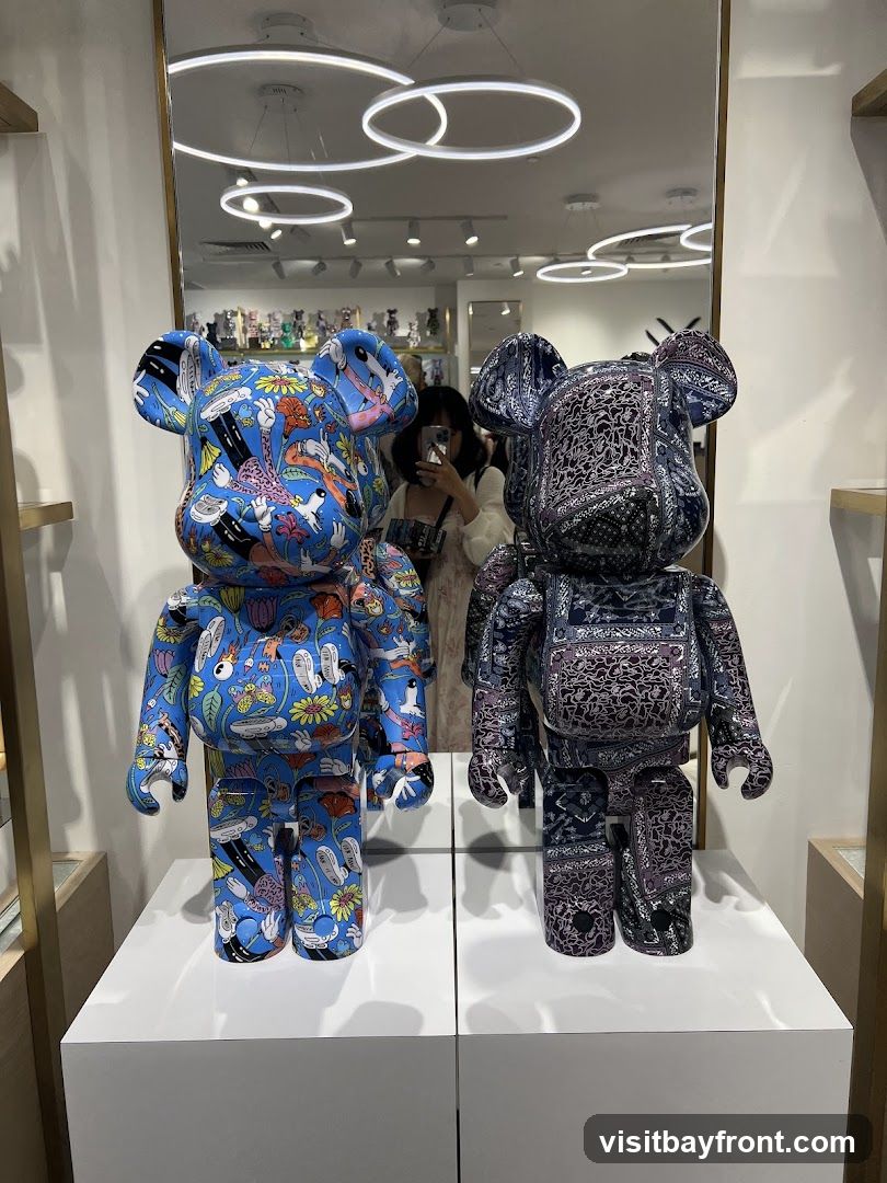Two large Bearbrick figures are displayed on white pedestals in front of a mirror. The figure on the left is blue with a colorful cartoon-like pattern featuring flowers, characters, and objects. The figure on the right is dark blue and black with a paisley-like pattern. The mirror reflects the interior of the store, including ceiling lights and shelves with smaller Bearbrick figures. A person is visible in the reflection, holding a phone.