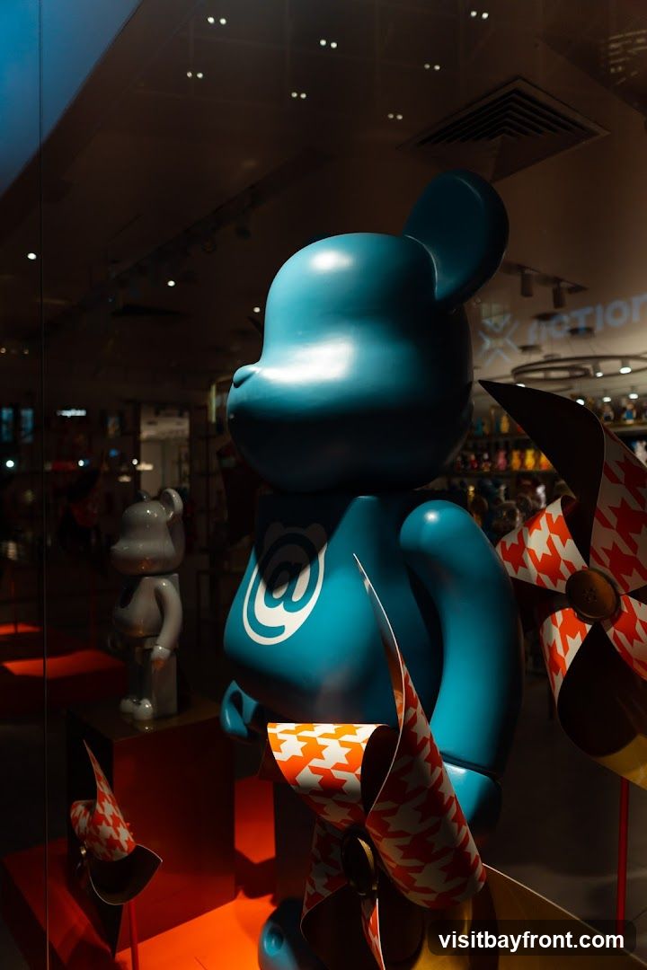 A display window features a large, teal-colored Bearbrick figure with an '@' symbol on its chest. To its left, a smaller, gray Bearbrick is visible. Several pinwheel-like decorations with a houndstooth pattern in orange and white are placed around the figures. The background shows a store interior with shelves stocked with various items and overhead lighting.