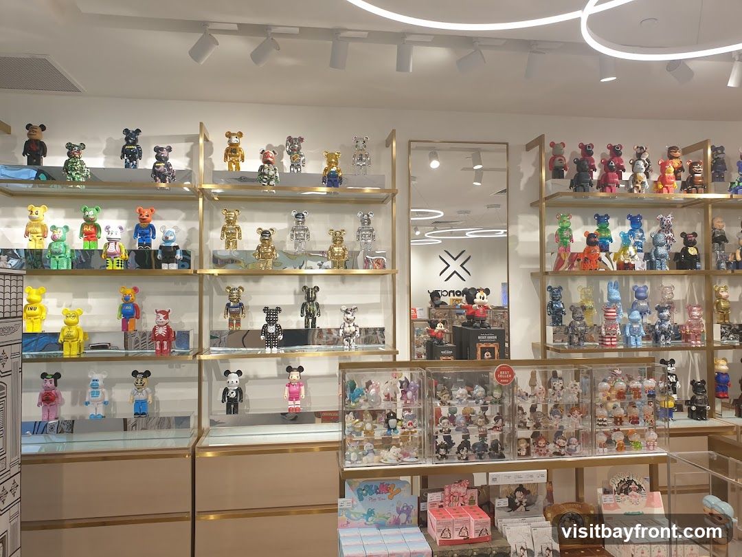 A well-lit interior view of a store showcasing a vast collection of collectible designer toys, primarily Bearbrick figures, arranged on multiple shelves. The toys are diverse in color, design, and size, with some featuring popular characters like Mickey Mouse. Display cases and boxes of merchandise are also visible, along with branded signage and decorative elements.